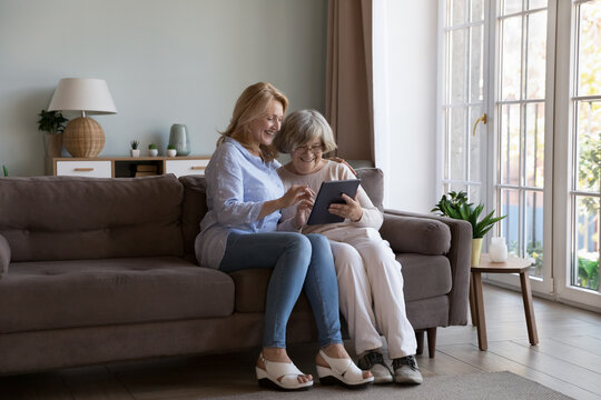Cheerful positive elderly mother and daughter making payment, video call on tablet computer. Caregiver teaching senior woman to use online app on for internet communication