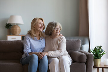 Happy dreamy middle aged daughter woman and old mom sitting close on home sofa, holding hands,...