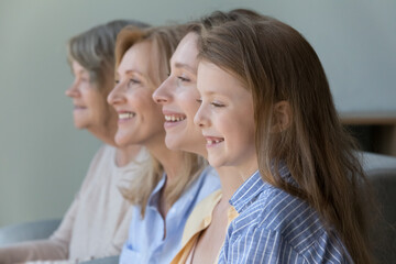 Cheerful cute kid girl standing near young mom, mature granny, old great grandmother, looking away, with tooth gap smile, thinking, laughing. Side portrait with four female generations, family dynasty