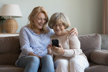 Happy nurse, caregiver helping cheerful elderly grandma woman to use online app on mobile phone. Senior mother and daughter sharing smartphone on home couch, making video call, enjoying communication