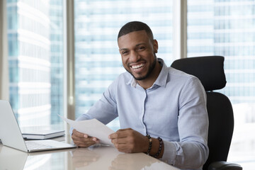 Happy African businessman, promoted employee, successful entrepreneur sit at workplace desk smile holds papers feels satisfied with sales growth, business success, posing in skyscraper office building