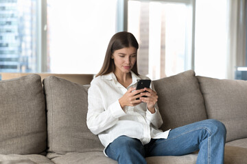 Obraz premium Young woman spend weekend on internet using modern smartphone, surfing web page, search information, makes call to friend or order using electronic web services resting on couch in modern living room