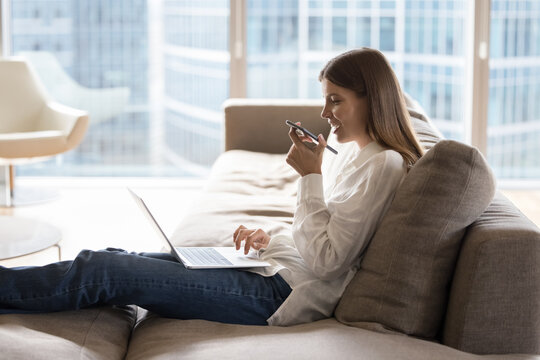 Attractive woman sits on sofa with laptop holds cellphone blab on speakerphone with friend while working, makes call to customer services, order goods at home use modern tech. AI tech usage, lifestyle