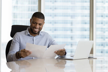 African business man in shirt holding sheets, read document paper report seated at office desk, company ceo doing paperwork feels satisfied with good work result, business growth and sales increase