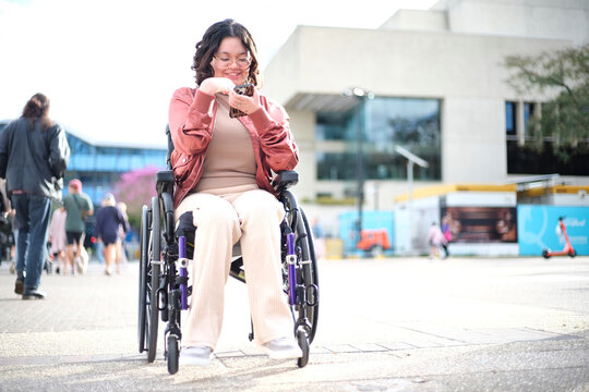 Smiling woman with a disability sitting in a wheelchair outside the city with mobile phone
