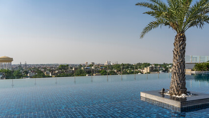 Fototapeta na wymiar Infinity pool on the roof. The blue tiled bottom is visible through the clear water. Palm tree against the blue sky. The city is visible in the distance. India