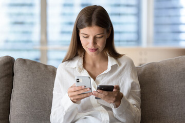 Woman holding two modern smart phones seated on couch, owner of new wireless gadget transfers data, contacts, files and photos from old model of device, compares software, use technology and back up