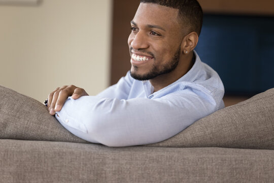 Handsome Millennial Single African Man Resting On Couch Staring Aside, Feels Satisfied, Enjoy Carefree Day Off, Relaxing On Pastime At Own Or Rented Modern Apartment. Homeowner Spend Leisure At Home