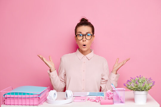 Amazed pretty brunette young woman with surprised expression spreads palms aside, has a lot of tasks during work day, sits at desktop with papers, isolated over pink background. Education and job