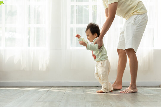 Adorable Asian Baby Toddler Learning To Walk With Dad Helping At Home. Little Baby Boy Looking In Front To Walking Step By Step And Father Supporting To Learning And Develop Skills Beside. 