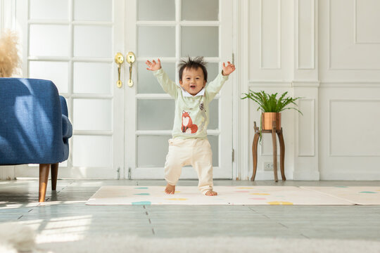 Adorable baby toddler learning to walking having fun and cheerful. Happy asian baby standing and raise hands up smile running and laughing at home.Good moment. Cute toddler looking at camera.