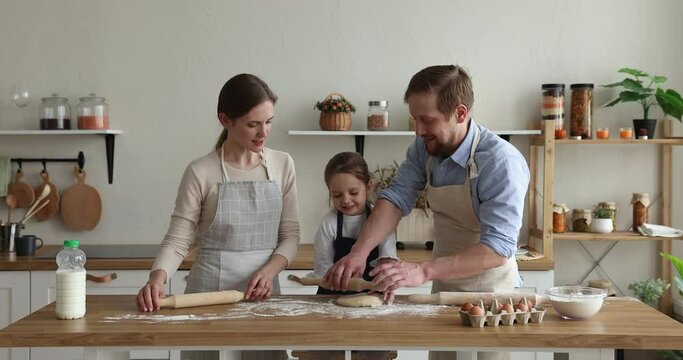 Loving parents teach daughter preparing cookies or family recipe pie, using rolling pin flattening dough for pastry enjoy cooking together wearing aprons standing and communicating in modern kitchen