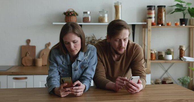 Millennial 35s couple absorbed in modern tech usage, holding two smart phones ignoring each other on leisure at home. Wireless technology addicted people, internet overuse problem, bad habit, phubbing
