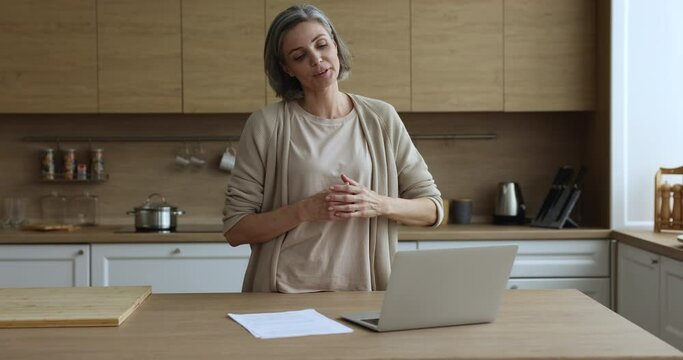 Attractive mature woman standing in kitchen, pass job interview on-line, staring at laptop screen makes speech, answers questions, working distantly, provide consultation to client by video conference