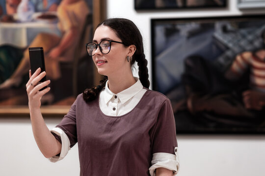 Cultural education. Portrait of caucasian pretty young woman wearing glasses visiting excursion in museum. Student using smartphone. Concept of exhibition in gallery