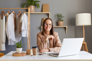 Indoor shot of attractive smiling woman wearing beige jacket sitting in front of laptop with clothing on hangers on background, working in showroom, being good mood.