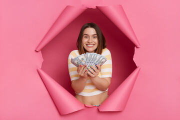 Indoor shot of delighted optimistic young girl wearing striped T-shirt breaking through paper hole of pink background, holding dollar banknotes, boasting her salary, winning lottery.