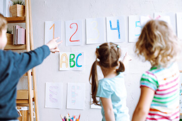 Children stand with back and learn english letters, alphabet and numbers on cards on wall. Kids point at colored numbers on sheets of paper by finger. Reading and counting, study in daycare centre - obrazy, fototapety, plakaty