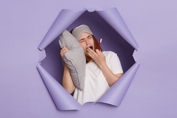 Image of sleepy young girl breaking through paper hole of purple background, holding toothbrush in...