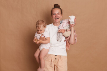 Indoor shot of smiling happy woman mother wearing white T- shirt standing with her little daughter...
