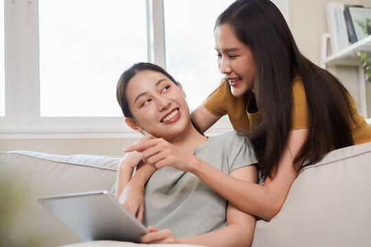 Happy lesbian, pleasure asian young two women, girl gay or close friend, couple love embrace, spending good time together, using tablet for entertainment on sofa at home. Activity of leisure, relax.