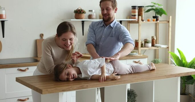 Cheerful playful family play together at home, loving parents fooling around holding rolling pin and flatten their daughter lying on counter top, make massage, having fun in modern domestic kitchen
