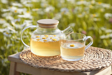 Transparent glass teapot with a mug of organic chamomile tea at placemat on nature background