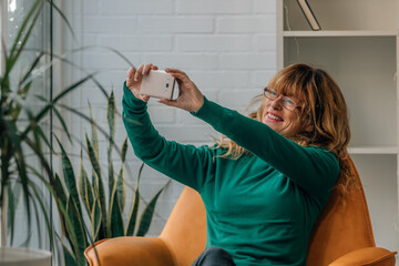 smiling middle aged woman with mobile phone at home making selfie