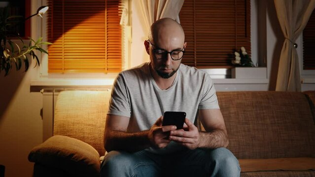Portrait of bald bearded Caucasian young man wearing glasses using smartphone. Evening cozy atmosphere. Concept of online communication.