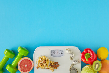 Slimming concept. Flat lay composition of scales with tape measure, dumbbells, vegetables and plate with nuts on blue background with copy space. Weight loss idea.