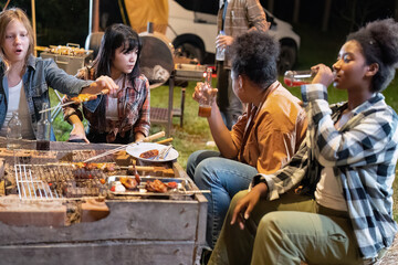 Group of family camper van sitting by fire grilling sausages and drinking dancing having dinner
