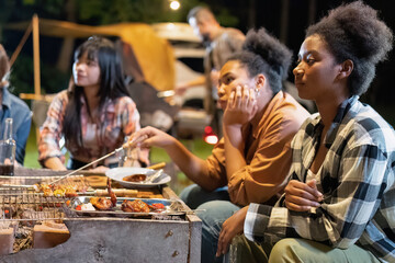 Group of family camper van sitting by fire grilling sausages and drinking dancing having dinner