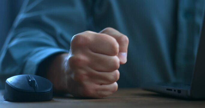 Close up aggressive man clicks computer mouse and furiously hits his fist on the table, emotional breakdown while working on laptop in the evening. Concepts of aggression at work at computer in office