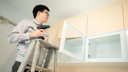 Asian male furniture assembler using electric drill screwdriver on cabinet hinge of glass panel. Interior construction worker man standing on ladder using screwdriving tools for furniture installation