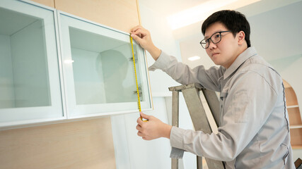 Home renovation or House remodeling concept. Asian male furniture assembler or Interior construction worker man standing on the ladder using tape measure on glass cabinet door panel.