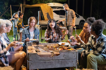 Group of family camper van sitting by fire grilling sausages and drinking dancing having dinner