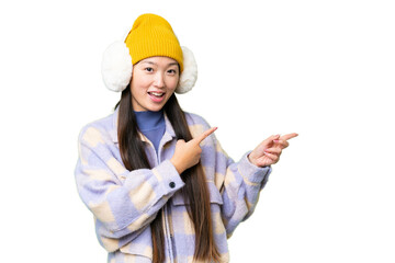 Young Asian woman wearing winter muffs over isolated chroma key background surprised and pointing side