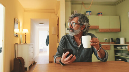 Elderly man drinks coffee from mug and uses mobile phone at home