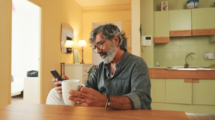 Elderly man drinks coffee from mug and uses mobile phone at home