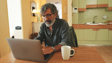 Elderly positive man typing on laptop sitting in home kitchen