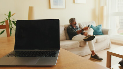 In the foreground, laptop and coffee mug are on the table, an elderly man is relaxing on the couch in the background. Soft focus