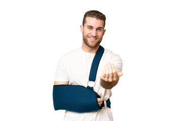 Young handsome blonde man with broken arm and wearing a sling over isolated chroma key background inviting to come with hand. Happy that you came