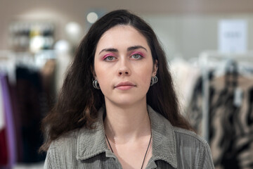 Shopaholic. Portrait of pretty sad young caucasian brunette woman with makeup. Defocused clothes store at background. Concept of psychology and mental health