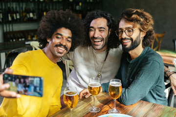 Young multiracial friends taking selfie with phone at table in restaurant bar. Three diverse young men having fun drinking beer together at brewery pub