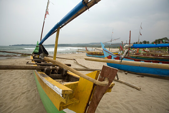 Narrow fishing boats pointed in the direct of the ocean sit on a sandy beach.