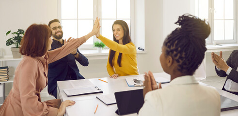 Two young women high five each other while mixed race coworkers are applauding. Team of happy business people celebrating results of successful teamwork during meeting in boardroom lit up by sun light - obrazy, fototapety, plakaty