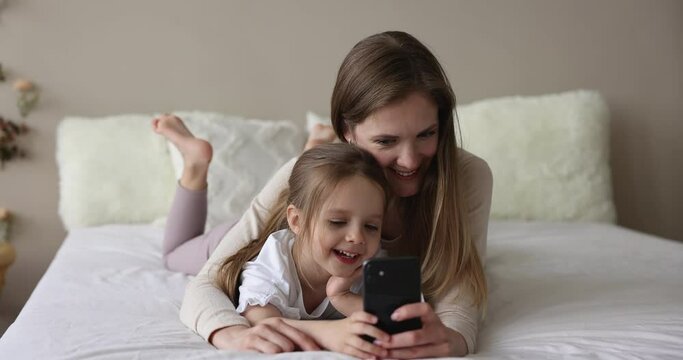 Parental control, leisure with modern wireless tech usage, lifestyle. Young woman and little cute daughter having fun lying on bed staring at cellphone screen laughing enjoy new mobile application