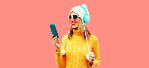 Winter portrait happy smiling woman looking at smartphone in headphones listening to music wearing yellow knitted sweater, white hat with pom pom, heart shaped sunglasses on pink background