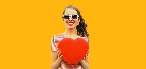 Portrait of happy smiling girl with red heart shaped balloon wearing sunglasses on yellow background
