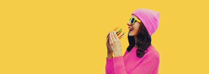 Portrait of stylish happy young woman with burger fast food wearing pink hat on yellow colorful background